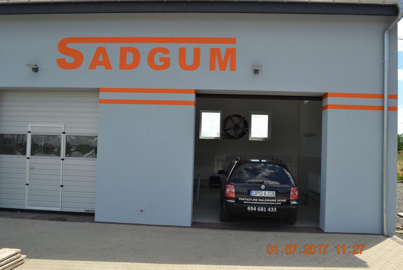 Szara elewacja budynku z pomarańczowym napisem SADGUM i pasami, garaż z szarą bramą i wjazdem, w którym stoi czarny samochód z napisem 'Fantazyjne malowanie ścian' na tylnej szybie.