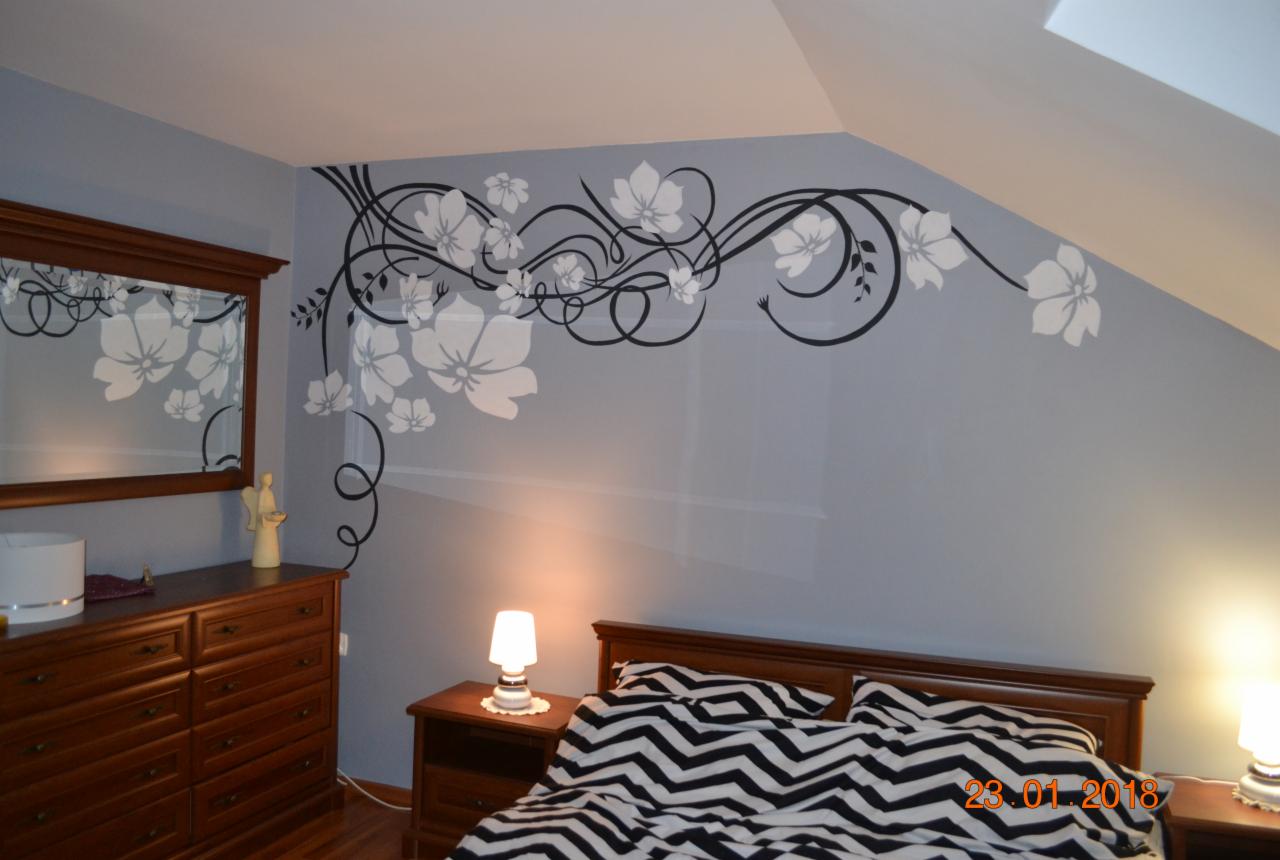 Pokój w Gościejowicach z szarą ścianą ozdobioną czarno-białym malarstwem artystycznym przedstawiającym kwiaty i pnącza, odbijające się w lustrze nad komodą.