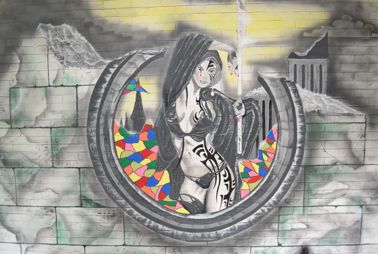 Artystyczny mural na ceglanej ścianie przedstawiający postać kobiety z tatuażami wewnątrz okręgu z kolorowymi elementami, trzymającej świetlny miecz, zarys zamku w tle.