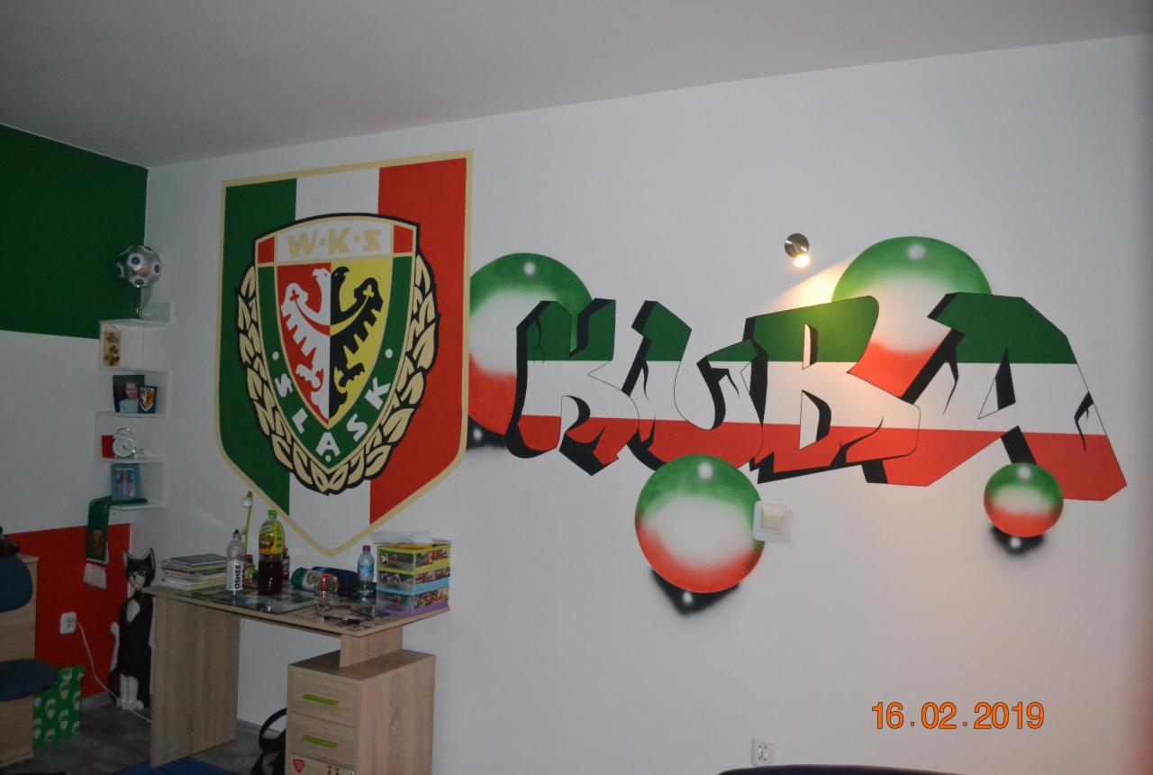 Pokój dziecięcy z muralem przedstawiającym herb klubu sportowego i graffiti w barwach klubowych na białej ścianie, biurko z zabawkami i półki z dekoracjami.