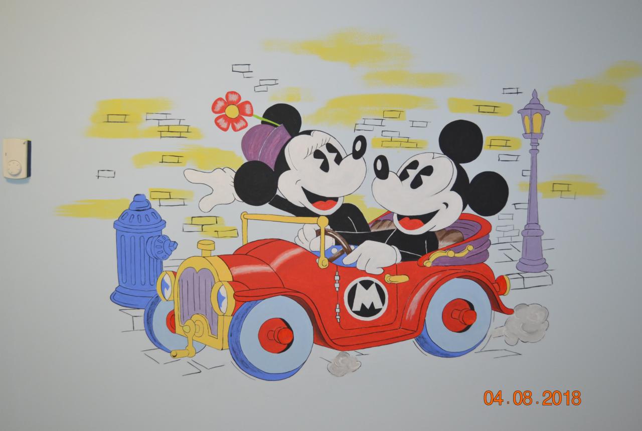 Ściana pokoju dziecięcego z ręcznie malowanym muralem przedstawiającym Myszki Miki w czerwonym samochodzie, na tle stylizowanego miejskiego krajobrazu z hydrantem i latarnią.