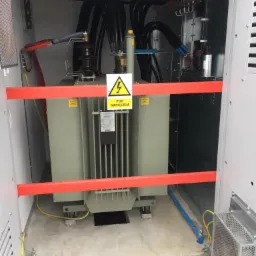 Transformator w obudowie z widocznym znakiem ostrzegawczym 'Pod napięciem', zabezpieczony czerwoną barierką, z grubymi kablami w czarnych osłonach.