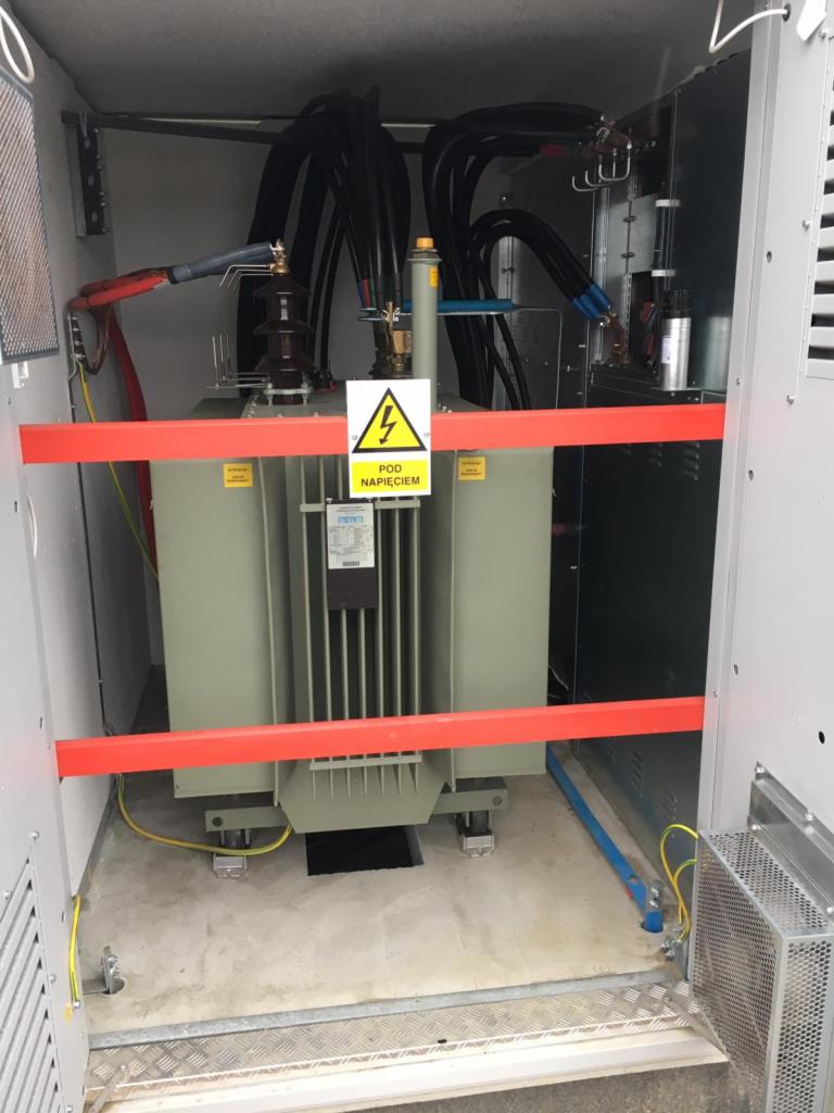 Transformator w obudowie z widocznym znakiem ostrzegawczym 'Pod napięciem', zabezpieczony czerwoną barierką, z grubymi kablami w czarnych osłonach.