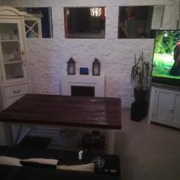 kris - Salon z dekoracyjną ścianą z białej cegły, imitacją kominka z lampionami, drewnianym stołem i szafką RTV z telewizorem.