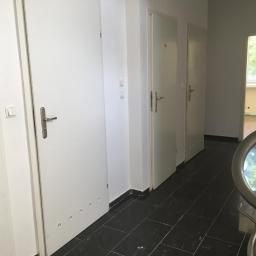 kris - Korytarz z trzema białymi drzwiami, czarną podłogą z płytek i metalową balustradą schodów po prawej stronie, widok z góry schodów.