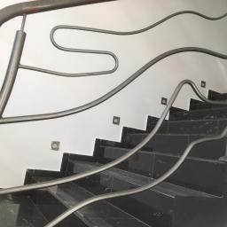 kris - Schody wyłożone czarnymi płytkami z marmurkowym wzorem w Wadowicach, z oryginalną, metalową balustradą o falistym kształcie i małymi, kwadratowymi lampkami ściennymi.