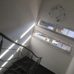 kris - Klatka schodowa z metalową balustradą, czarnymi stopniami i białymi ścianami oświetlona promieniami słońca wpadającymi przez okna.