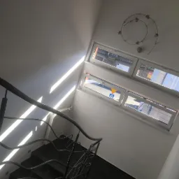 Klatka schodowa z metalową balustradą, czarnymi stopniami i białymi ścianami oświetlona promieniami słońca wpadającymi przez okna.