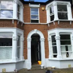 Odnowiona fasada domu z czerwonej cegły z białymi elementami dekoracyjnymi okien i wejścia, widoczne puszki farby i drobne zabrudzenia na chodniku.