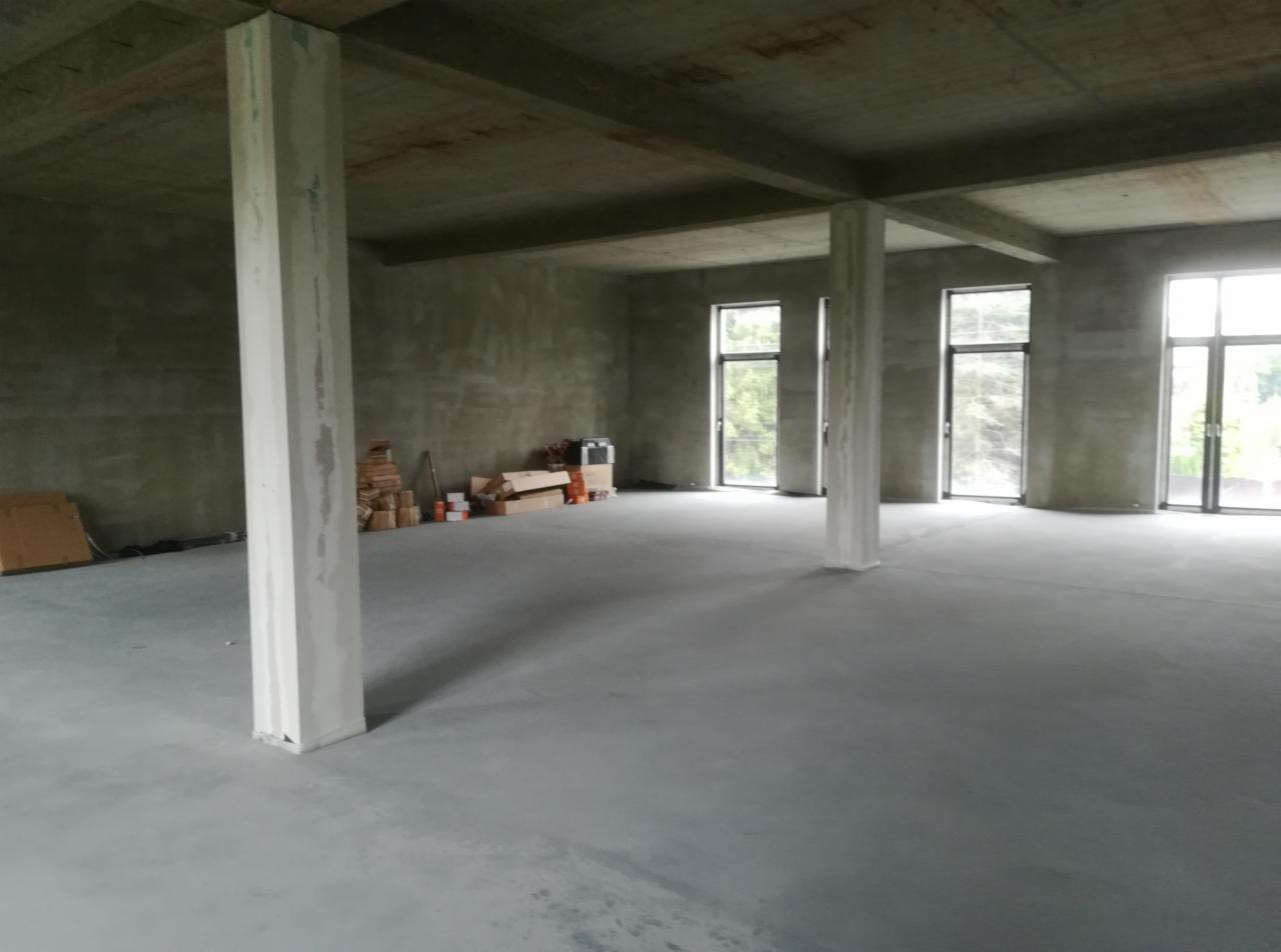 Surowe wnętrze w stanie deweloperskim z betonowymi ścianami, kolumnami i wylewką, przygotowane do prac wykończeniowych, z widocznymi materiałami budowlanymi w tle.