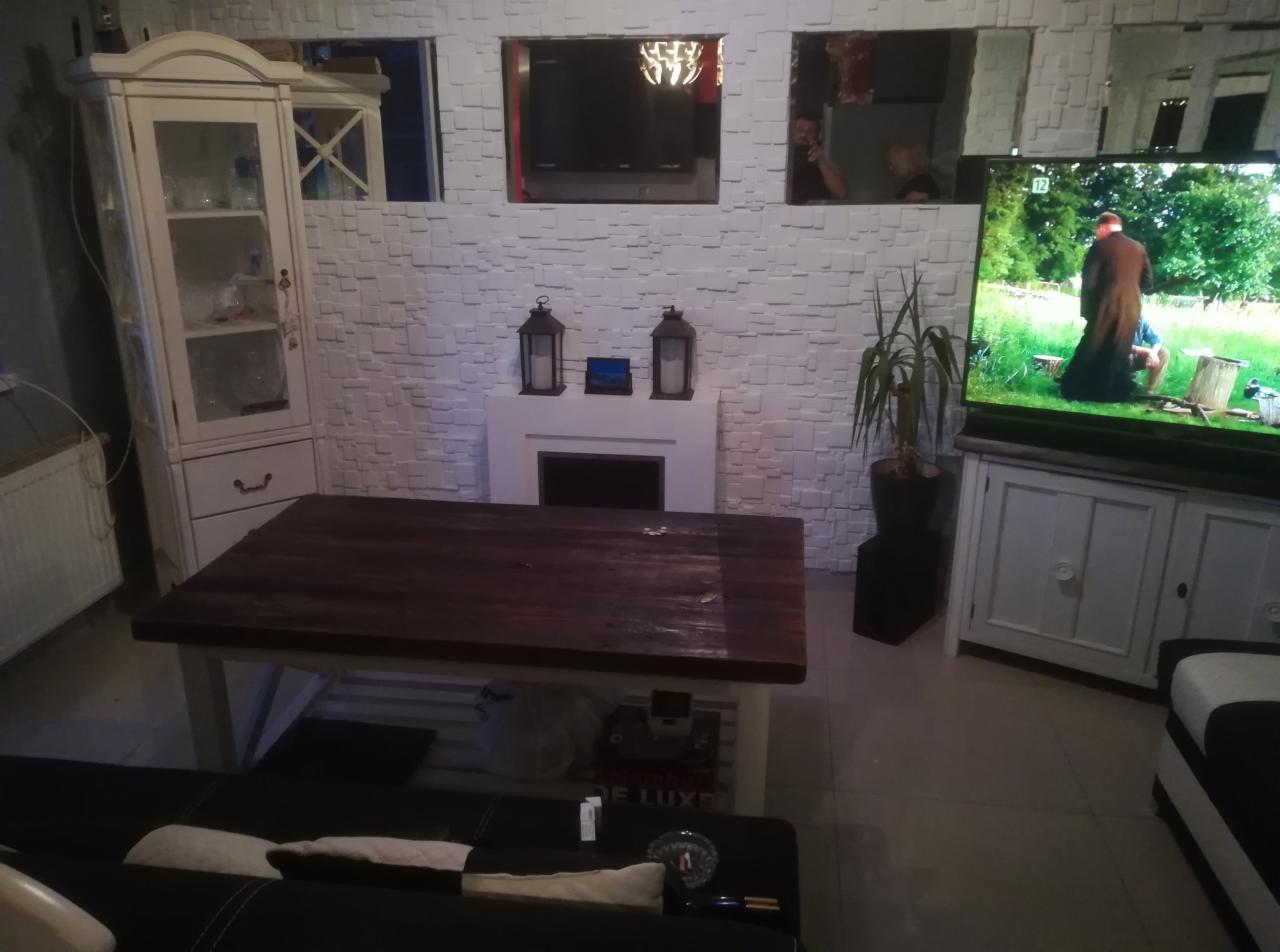 Salon z dekoracyjną ścianą z białej cegły, imitacją kominka z lampionami, drewnianym stołem i szafką RTV z telewizorem.