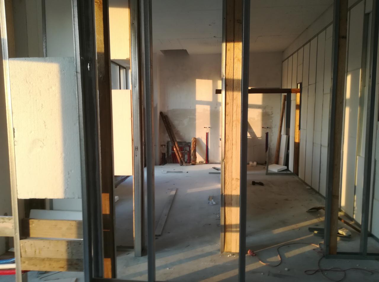 Wnętrze w trakcie montażu ścian działowych z płyt kartonowo-gipsowych, widoczne metalowe profile i fragmenty płyt, surowe ściany w tle, wpadające promienie słoneczne.