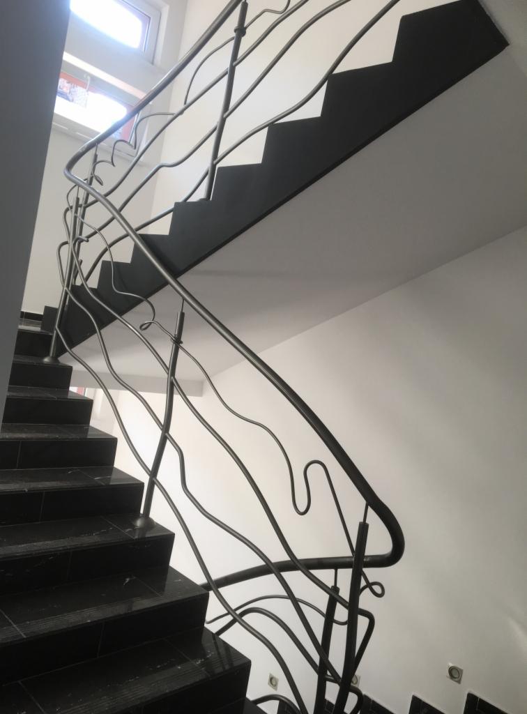 Czarne, marmurowe schody z oryginalną, giętą, metalową balustradą na tle białej ściany i sufitu.