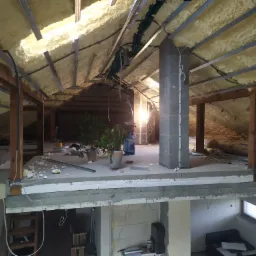 Poddasze w trakcie ocieplania wełną mineralną, widoczne krokwie, profile metalowe, fragment instalacji elektrycznej i wentylacyjnej oraz rośliny doniczkowe na podłodze