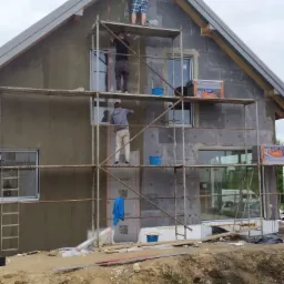 Ocieplanie elewacji domu jednorodzinnego w Kocku: Trzech pracowników na rusztowaniu aplikuje tynk na ocieplone ściany, widoczne paczki styropianu, niezagospodarowany teren wokół budynku.