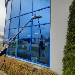 Czyszczenie niebieskich okien budynku komercyjnego za pomocą szczotki na długim wysięgniku, w tle widoczny budynek i zieleń.