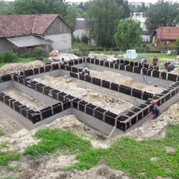 Budowa fundamentów domu jednorodzinnego z użyciem szalunków i bloczków betonowych, widoczni pracownicy budowlani, w tle domy i drzewa.