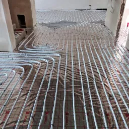 Rozłożona instalacja ogrzewania podłogowego na folii izolacyjnej z widocznymi rurami grzewczymi mocowanymi do siatki, w surowym stanie wykończenia wnętrza.