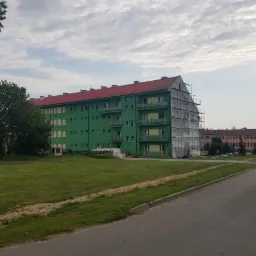 Wielopiętrowy budynek z czerwoną dachówką, częściowo pokryty rusztowaniami w trakcie prac elewacyjnych, z widocznym fragmentem ocieplonej ściany i stertą materiałów budowlanych na trawniku.