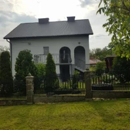 Dwupiętrowy dom z szarą elewacją, czarnym dachem i kutą balustradą balkonu, otoczony zielonym trawnikiem i ogrodzeniem z kamiennymi słupkami.