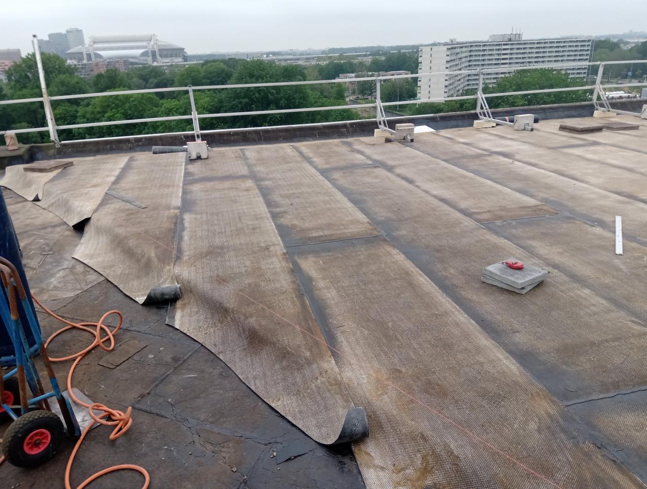 Rozwinięte pasy papy na płaskim dachu z widoczną strukturą siatki, częściowo przyklejone, z wózkiem transportowym i narzędziami w tle widok na miasto.