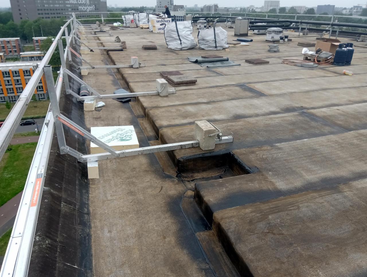 Płaski dach pokryty papą bitumiczną z widocznymi elementami zabezpieczeń krawędzi, workami z materiałem i narzędziami, w tle zabudowa miejska.