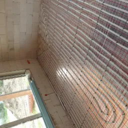 Białe rury ogrzewania podłogowego ułożone w pętle na czerwonej folii z siatką, widok z góry, surowy stan ścian z białych pustaków, okno z widokiem na zewnątrz.