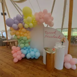 Dekoracja balonowa w pastelowych kolorach, tworząca łuk przy białej ściance z napisem 'Roczek Marlenki', ustawiona w namiocie na drewnianej podłodze.