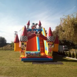 Kolorowy dmuchany zamek z motywem Myszki Miki, ustawiony na trawniku w Oświęcimiu, z widocznymi elementami dekoracyjnymi i schodami. Na tle drzewa i błękitne niebo.