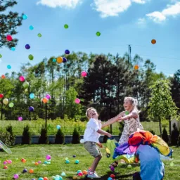 Dzieci bawią się kolorowymi piłeczkami na trawie pod błękitnym niebem, trzymając barwną chustę animacyjną.