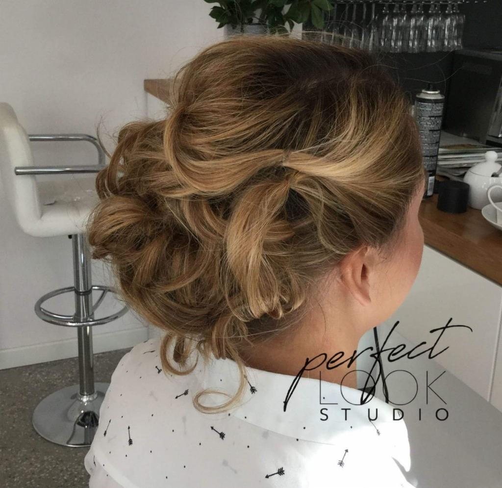 Upięcie włosów w stylu messy bun z blond pasemkami, widok z tyłu, w tle krzesło barowe i fragment blatu z akcesoriami.