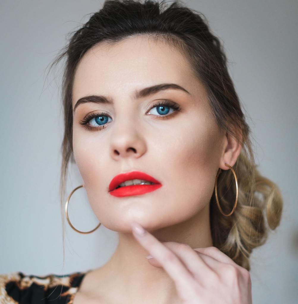 Portret kobiety z intensywnie czerwoną pomadką, podkreślonymi brwiami i niebieskimi oczami, złote kolczyki koła, palec wskazujący na brodę.