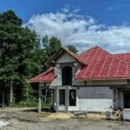 Niedokończony dom jednorodzinny z czerwoną konstrukcją dachu, widok z zewnątrz na tle lasu i błękitnego nieba z chmurami.