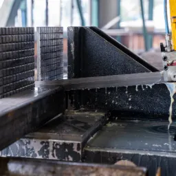 Piła taśmowa przecina metalowy profil, chłodziwo spływa po ciętym materiale, widoczne imadło i elementy mocujące.