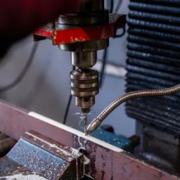 Wiertło frezarki CNC w trakcie obróbki metalowego profilu, widoczne chłodziwo i wióry, oświetlenie warsztatowe.