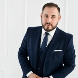 Mężczyzna w granatowym garniturze z poszetką, stojący na tle białej ściany z panelami. Uśmiech, zadbana broda i włosy. Profesjonalny wizerunek.