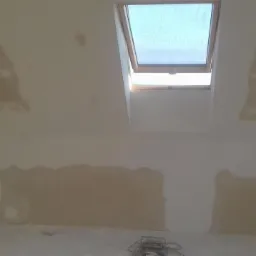 Poddasze w trakcie prac wykończeniowych, widoczne fragmenty ściany i sufitu z nałożoną warstwą wyrównującą, przygotowane do dalszych prac, okno dachowe w tle.