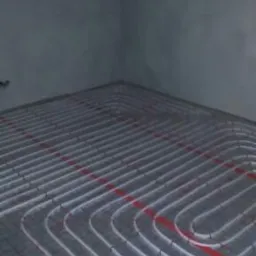 Montaż ogrzewania podłogowego w nowym budynku w Jędrzejowie; widoczne białe rury ułożone w pętle na szarej podłodze, przymocowane do czerwonej siatki.