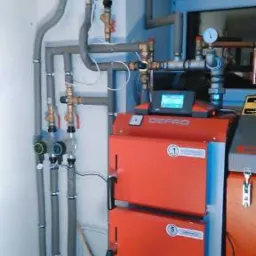 Czerwony piec CO marki DEFRO z podłączonymi rurami instalacji grzewczej, widoczne zawory i manometry, szare rury prowadzone wzdłuż ściany.