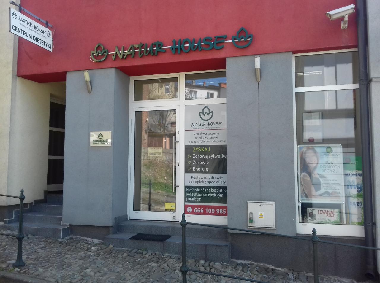 Fasada budynku Natur House z szyldem i plakatem reklamowym, oferującego konsultacje dietetyczne i poprawę sylwetki, widoczny numer telefonu.