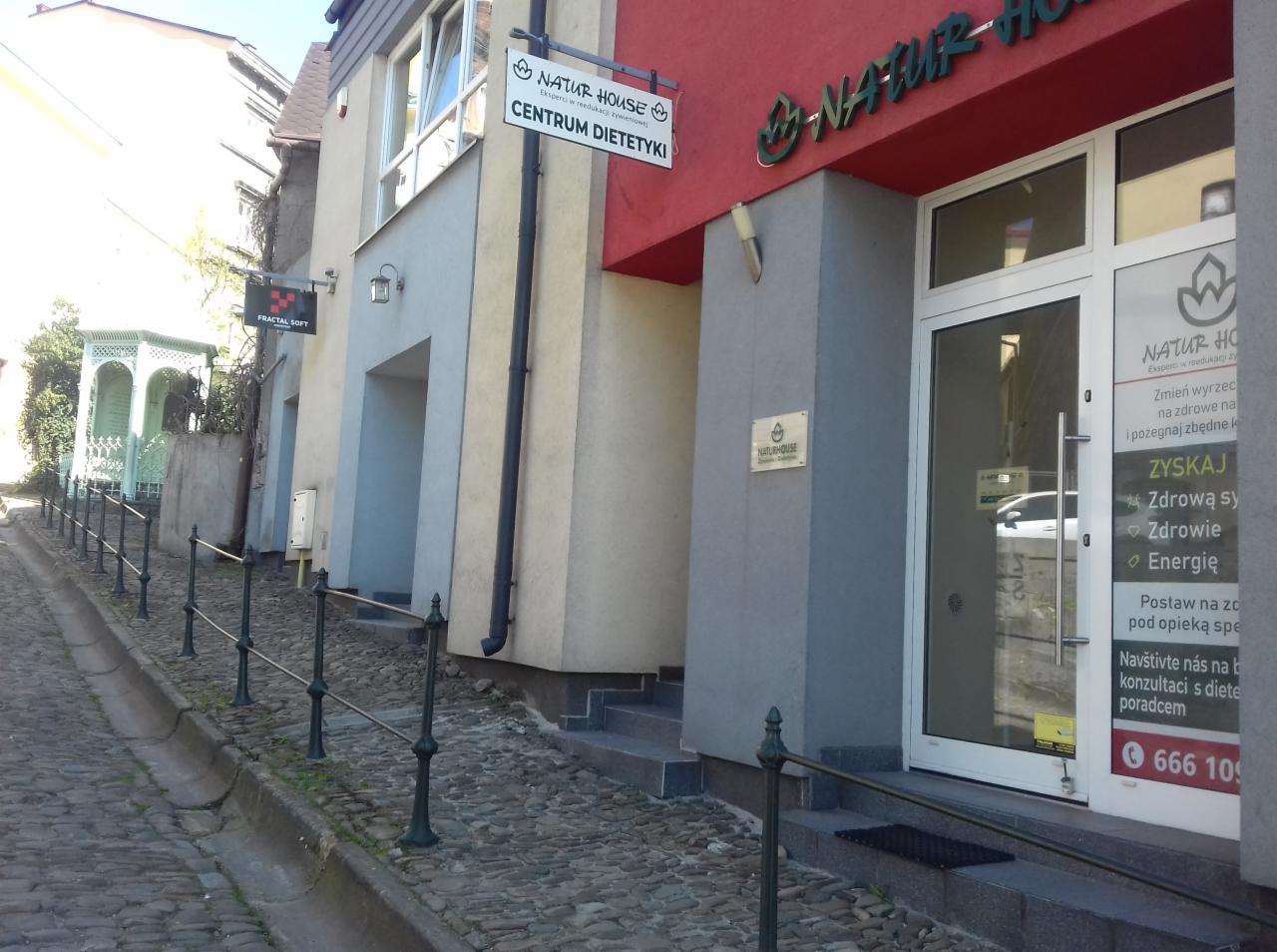 Wejście do Centrum Dietetyki Natur House z szyldem i reklamą na drzwiach, widok z brukowanej uliczki pod górę.