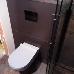 Nowoczesna, podwieszana toaleta w łazience z ciemnymi, matowymi płytkami, widoczna częściowo kabina prysznicowa i niedokończone wykończenie ściany po lewej stronie.