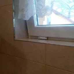 Beżowe płytki okalające okno z białą ramą i koronkową firanką, widok na zewnątrz