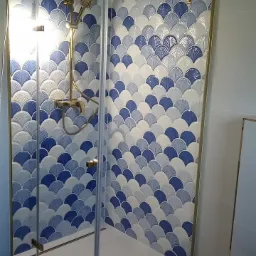 Kabina prysznicowa z mozaiką w odcieniach błękitu i bieli, ułożoną w kształt wachlarzy. Złota armatura i rama kabiny dodają elegancji.