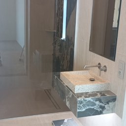 Usługi remontowe Glazurnik - Nowoczesna łazienka z prysznicem walk-in, kamienną umywalką na podświetlanej szafce, lustrem i ścianą z ciemnych płytek imitujących marmur. Na podłodze pasujący kamienny blok.