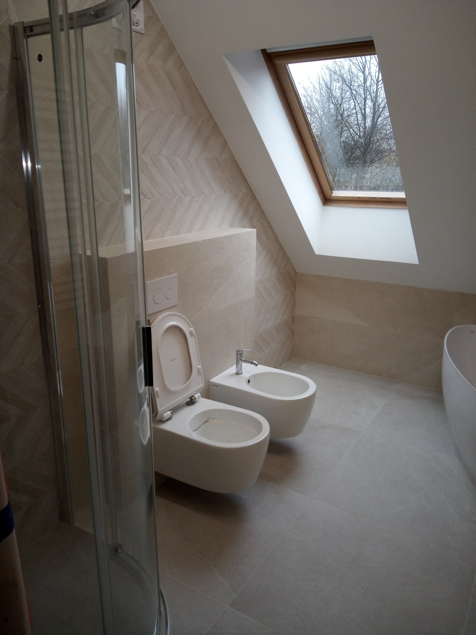 Jasna łazienka na poddaszu z oknem dachowym, kabiną prysznicową, wiszącą toaletą z deską i bidetem, wykończona płytkami w odcieniach beżu z delikatnym wzorem jodełki na ścianach, widok na drzewa...