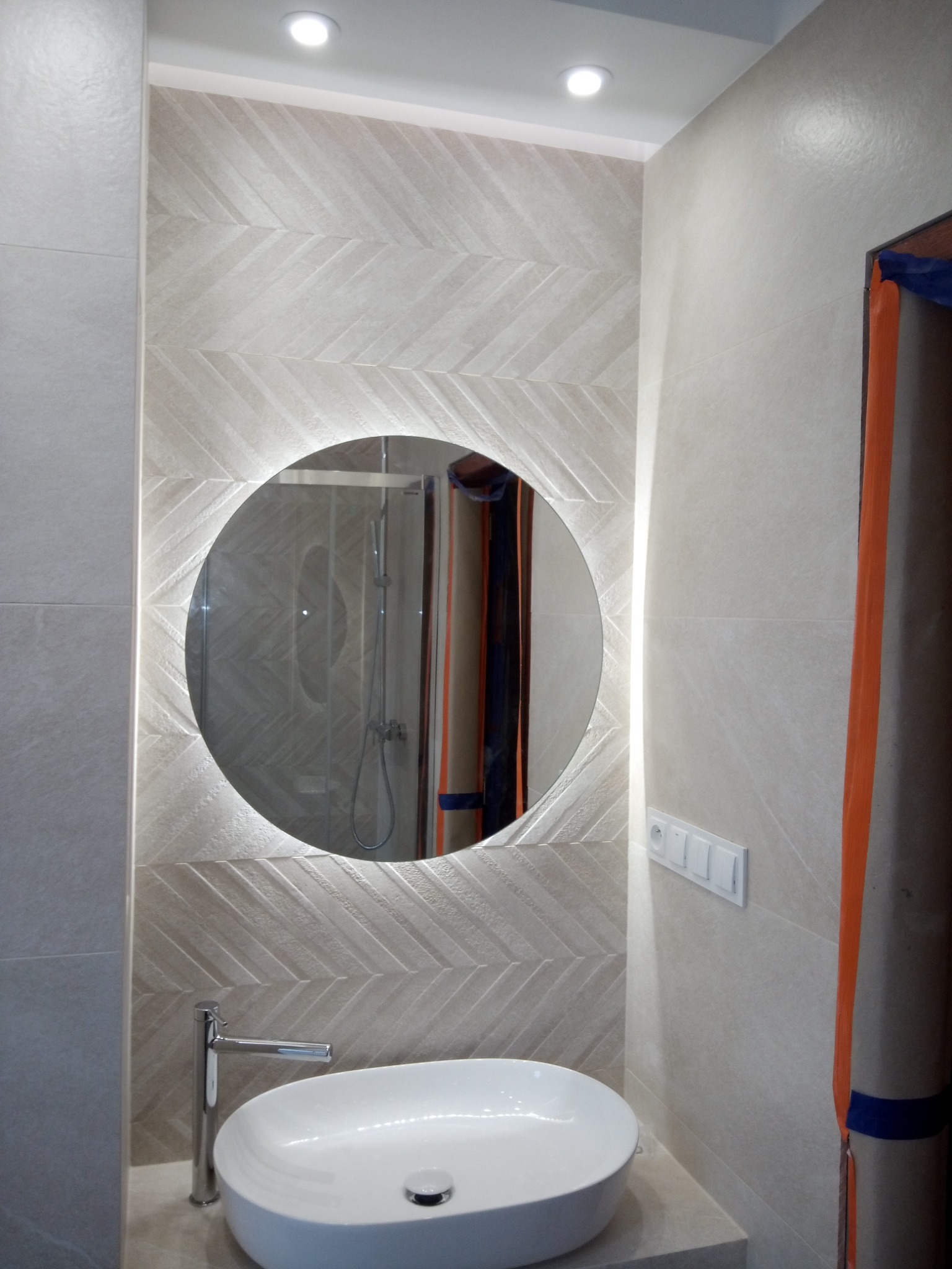 Wykończona łazienka w Jaśle: okrągłe lustro z podświetleniem LED nad białą, owalną umywalką nablatową i chromowaną baterią, ściany wyłożone płytkami w odcieniach beżu z wzorem jodełki.