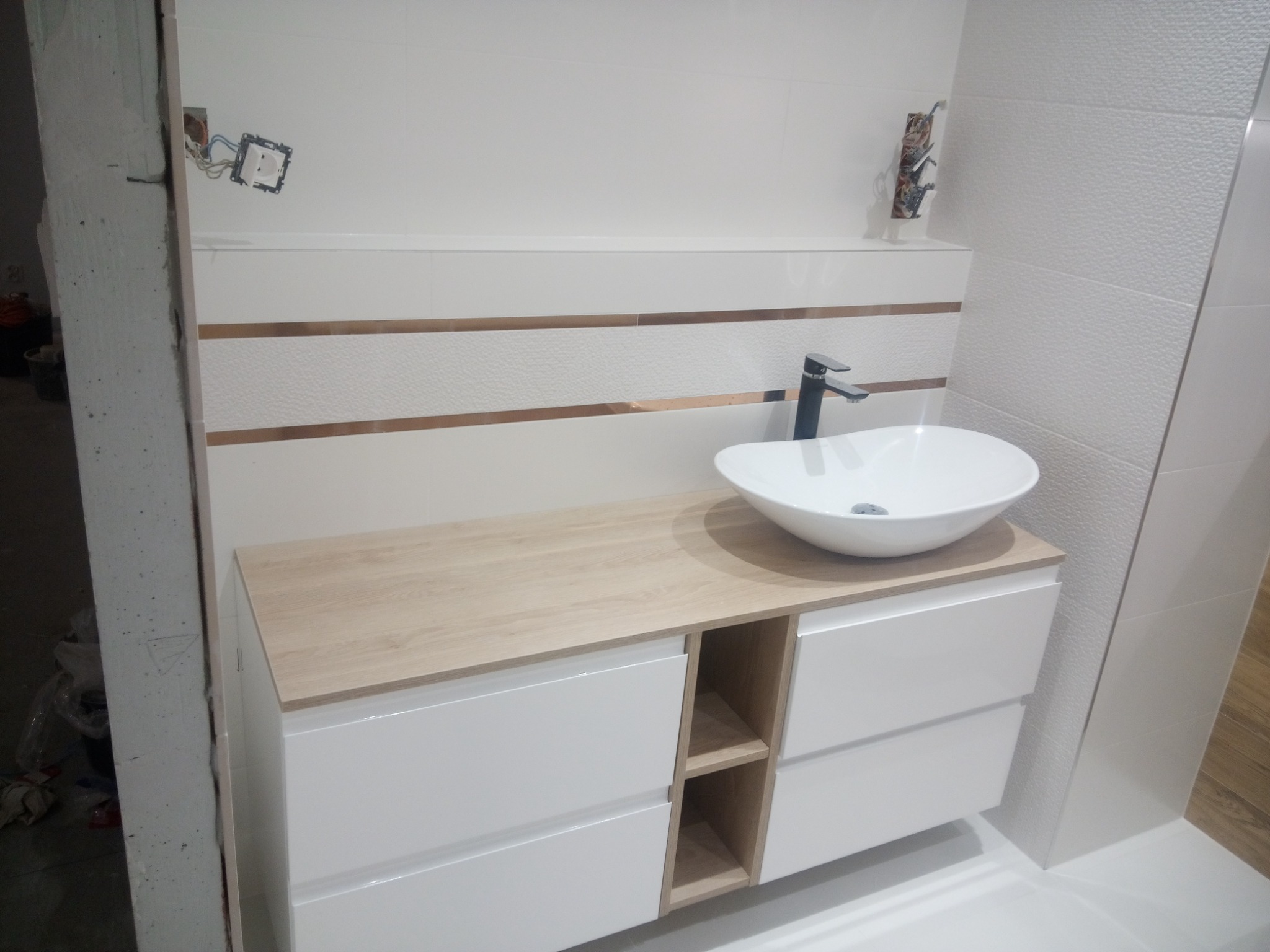 Nowoczesna łazienka w trakcie wykańczania: białe płytki z poziomymi, złotymi paskami, biała umywalka nablatowa na szafce z jasnego drewna, widoczne niedokończone gniazdko elektryczne.