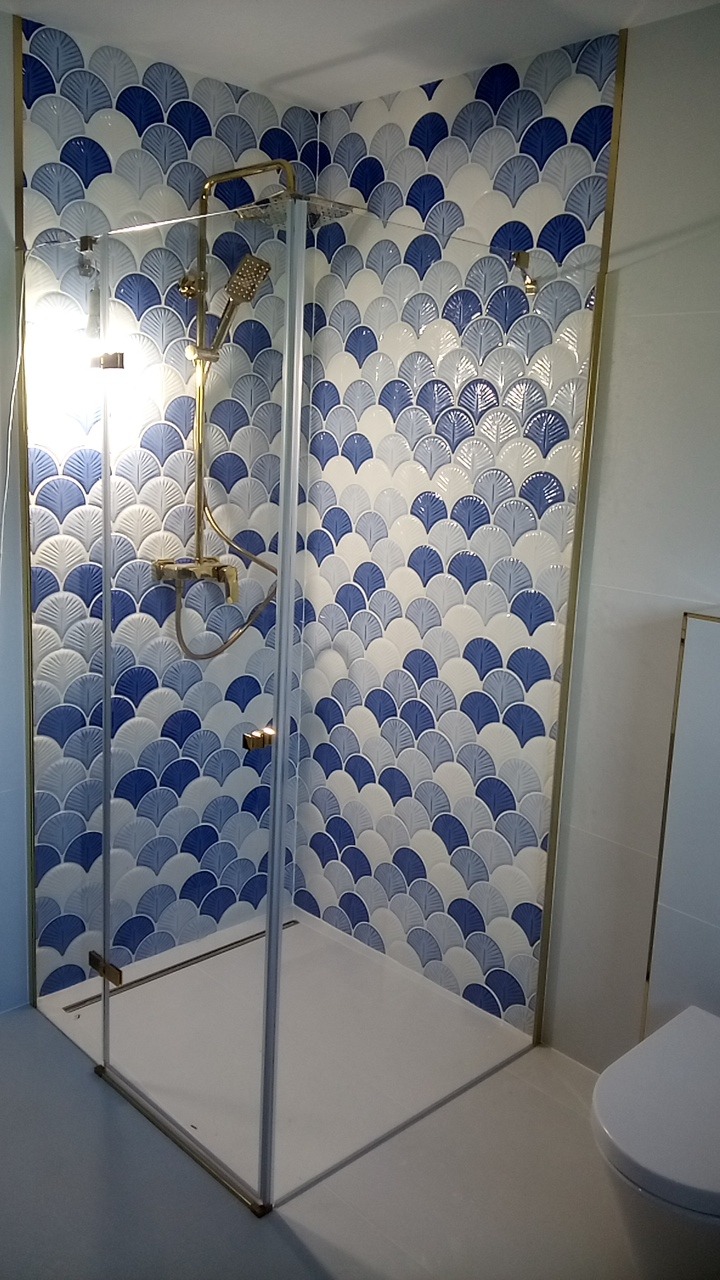 Kabina prysznicowa z mozaiką w odcieniach błękitu i bieli, ułożoną w kształt wachlarzy. Złota armatura i rama kabiny dodają elegancji.