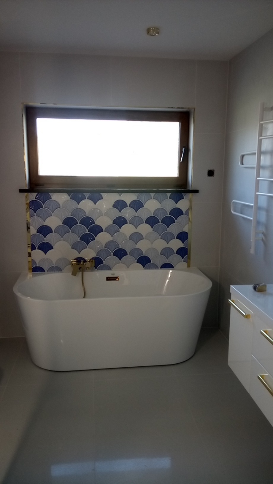 Biała, wolnostojąca wanna pod oknem z mozaiką w łazience z szarymi płytkami i białymi szafkami z złotymi uchwytami. Nowoczesny styl.